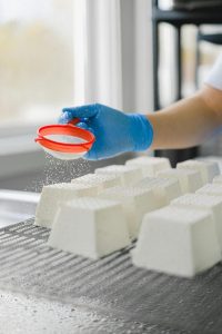 Close-up of cheese being sprinkled with a sieve in a production facility, highlighting hygiene practices.