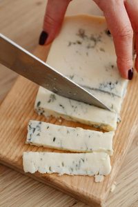 Close-up of a hand slicing creamy Gorgonzola cheese with a knife.